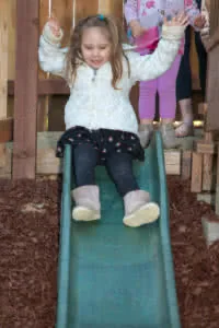 Young child sliding down a green playground slide with arms raised, wearing white jacket and pink shoes, other children visible in background
