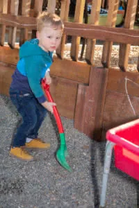 Toddler in blue jacket and jeans holding a red and green shovel while standing on gravel near a wooden fence