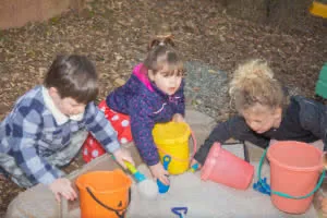 Three young children playing with colored buckets and shovels in sandbox outdoors