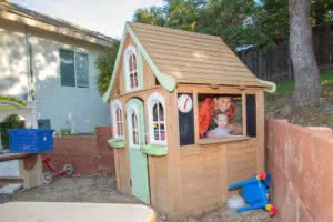 Two children playing inside a wooden playhouse with a peaked roof and green trim in a backyard setting.