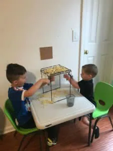 Two young children sitting at a white table with green chairs, playing with straw pieces in a black wire tray during a sensory activity.