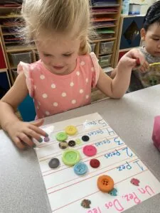 Young child with blonde hair wearing pink polka dot shirt pointing at educational worksheet with colored buttons sorted by size at classroom table