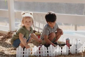 Two young children playing in indoor sandbox with toy farm animals and white fence