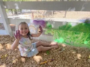 Young child sitting on mulch with hands covered in purple and green paint, smiling at window with colorful paint streaks