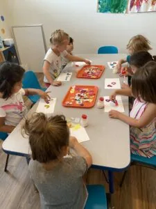 Children sit around a table doing art activities with paint and paper in a classroom setting.