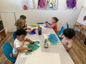 Five young children painting with watercolors at a table in an art studio with finished artwork displayed on the wall behind them.