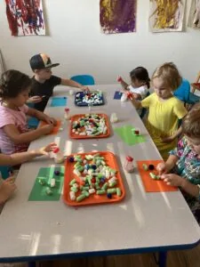 Children sitting at tables with colorful foam pom-poms in orange trays during a classroom craft activity