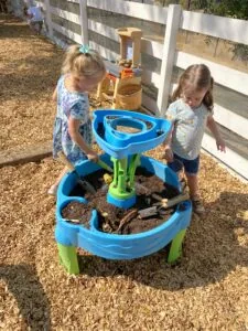Two young children using a blue and green water table with multiple basins in an outdoor play area with white fencing and wood chips.