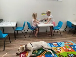 Two young children sit at a white table with blue chairs in a classroom, while a light-colored dog lies on an educational mat on the wooden floor.
