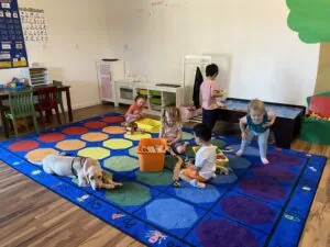 Young children playing on a colorful educational floor mat in a classroom with a dog, toy kitchen, and learning materials visible.