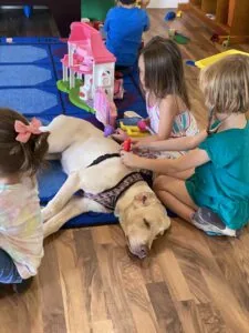 Children petting and playing with a light-colored dog on a playroom floor surrounded by toys and colorful play structures.