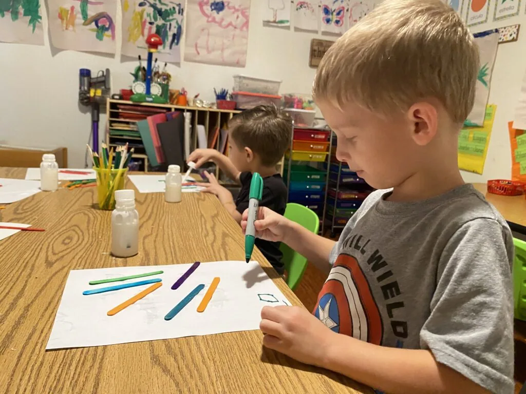 Two children drawing with markers at a wooden table in a classroom with art supplies and student artwork on walls.